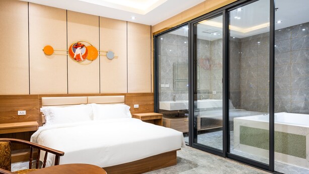 A modern hotel room with a large bed, a wooden headboard, a nightstand, a chair, and a glass-enclosed bathroom with a bathtub. - Home me Hotel (Makati)