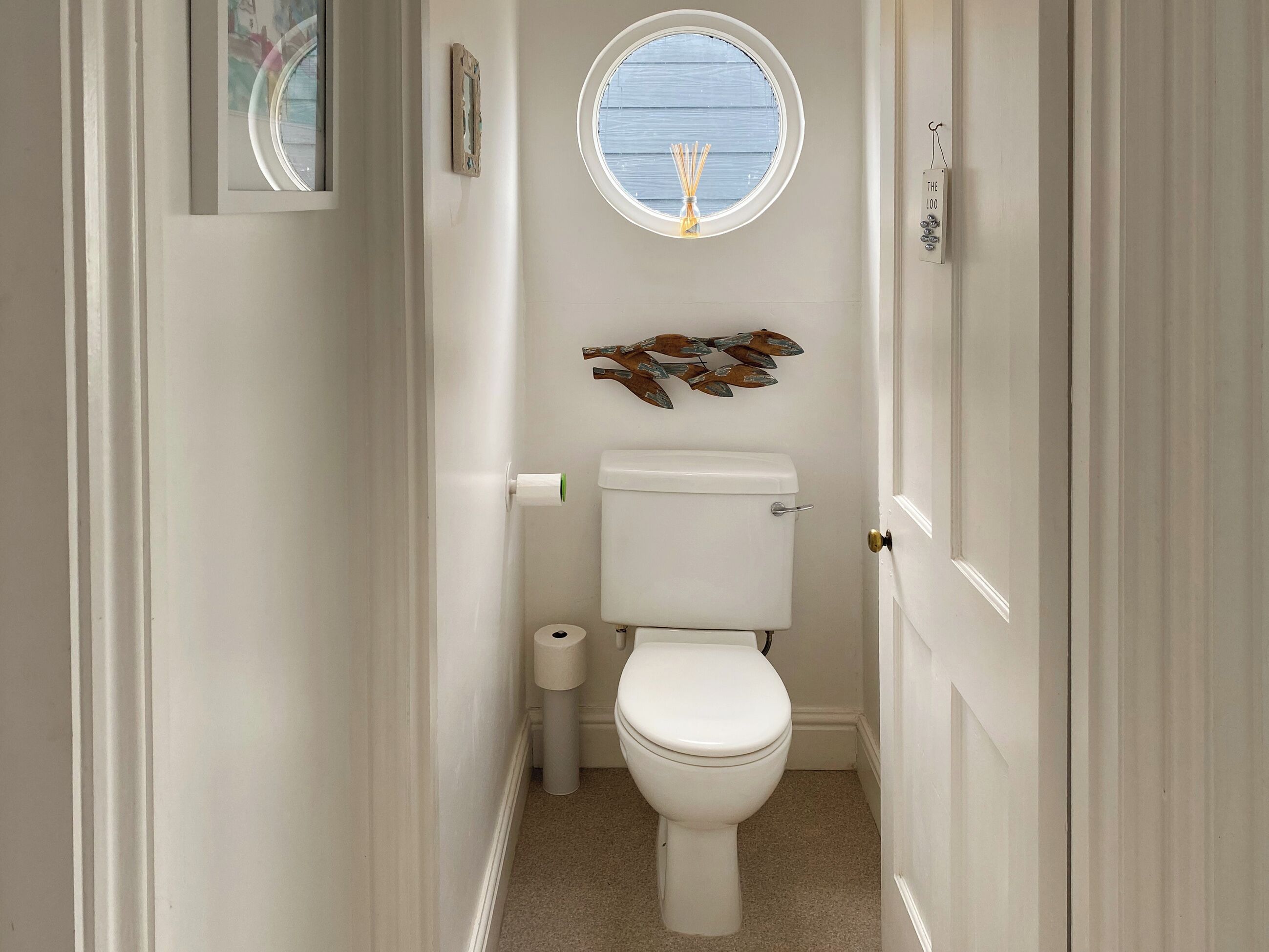 A narrow bathroom with a toilet, a small window, and a decorative wall piece.