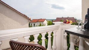 A balcony with a white balustrade, a table with a bowl of fruit and wine glasses, and a view of houses and greenery. - Two bedroom apartment for 3 person (Jadranovo)