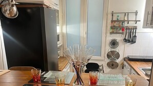 A kitchen with a wooden island, two chairs, a refrigerator, and a window with frosted glass panels. - Montmartre-appartement Climatisé -3min Sacré-cœur (Paris)