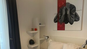 A neatly made bed with white linens, a bedside table with a lamp, and a wall-mounted shelf with a few items. - Montmartre-appartement Climatisé -3min Sacré-cœur (Paris)