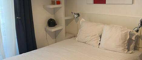 A neatly made bed with white linens, a bedside table with a lamp, and a wall-mounted shelf with a few items.