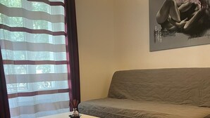 A room with a wooden floor, a grey sofa, a white table with a vase, and a window with striped curtains. - Montmartre-appartement Climatisé -3min Sacré-cœur (Paris)