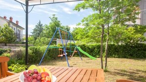 An outdoor wooden table with a bowl of fruit, a swing set, and a slide in the background.