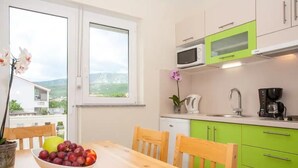 A kitchen with a wooden table, chairs, a bowl of fruit, a microwave, a coffee maker, and a view of the outdoors.
