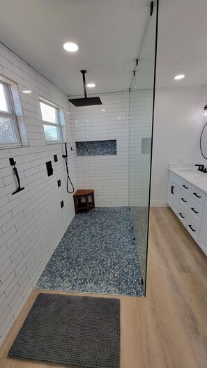 Une salle de bain moderne avec des murs recouverts de carreaux blancs, une douche dotée d’une porte en verre, un plancher en bois et un petit tabouret en bois.