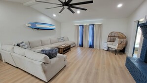 A modern living room with a sofa, a coffee table, a wicker chair, a ceiling fan, and a wall-mounted surfboard.
