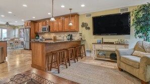 A spacious kitchen with wooden cabinets, a central island with stools, a large flat-screen TV, and a cozy seating area with a sofa and a plant.