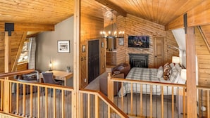 Een gezellig interieur van een blokhut met een slaapkamer op een entresol, een open haard, een bureau met stoel en een eethoek.