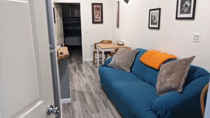 Une pièce avec un canapé bleu, deux coussins, une petite table sur laquelle se trouve un bol, et une chaise. Une porte donne accès à une autre pièce.