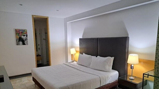 A hotel room with a large bed, two bedside tables with lamps, a wooden headboard, a painting on the wall, and a mirror on the right side. - AW HOTEL LUXOR VERSALLES (Cali)
