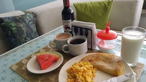 A breakfast setting with a plate of scrambled eggs, a slice of toast, a slice of watermelon, a bottle of wine, a cup of coffee, a glass of milk, and a red drink on a table with a placemat. - AW HOTEL LUXOR VERSALLES (Cali)
