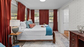 A bedroom with a bed, a bedside table with a lamp, a dresser, and a window with curtains.
