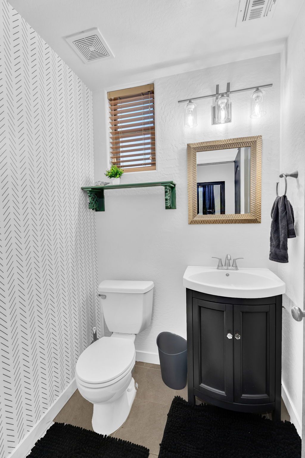 Ein Badezimmer mit einer weißen Toilette, einem schwarzen Waschtisch mit Spiegel und einem kleinen Fenster mit Jalousien.