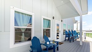 Un área de estar al aire libre techada con sillas Adirondack azules y una mesa, paredes blancas y ventanas grandes con cortinas.