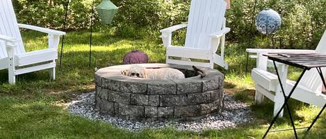 Un hogar circular de piedra rodeado de sillas Adirondack blancas en un área boscosa.