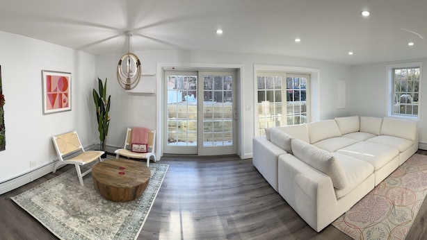 Ein geräumiges Wohnzimmer mit einem großen weißen Sofa, einem Holztisch und einem gemusterten Teppich. Zwei Sessel, ein großes Fenster und eine dekorative Wandgestaltung sind ebenfalls vorhanden.