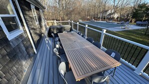 Eine Holzterrasse mit Tisch und Stühlen, einem überdachten Bereich und Blick auf einen Garten mit Bäumen und Rasen.