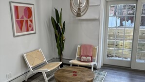 Ein geräumiges Zimmer mit einem runden Holztischchen, zwei weißen Stühlen, einer großen Topfpflanze, einem gerahmten abstrakten Kunstwerk und Blick durch eine Glastür.