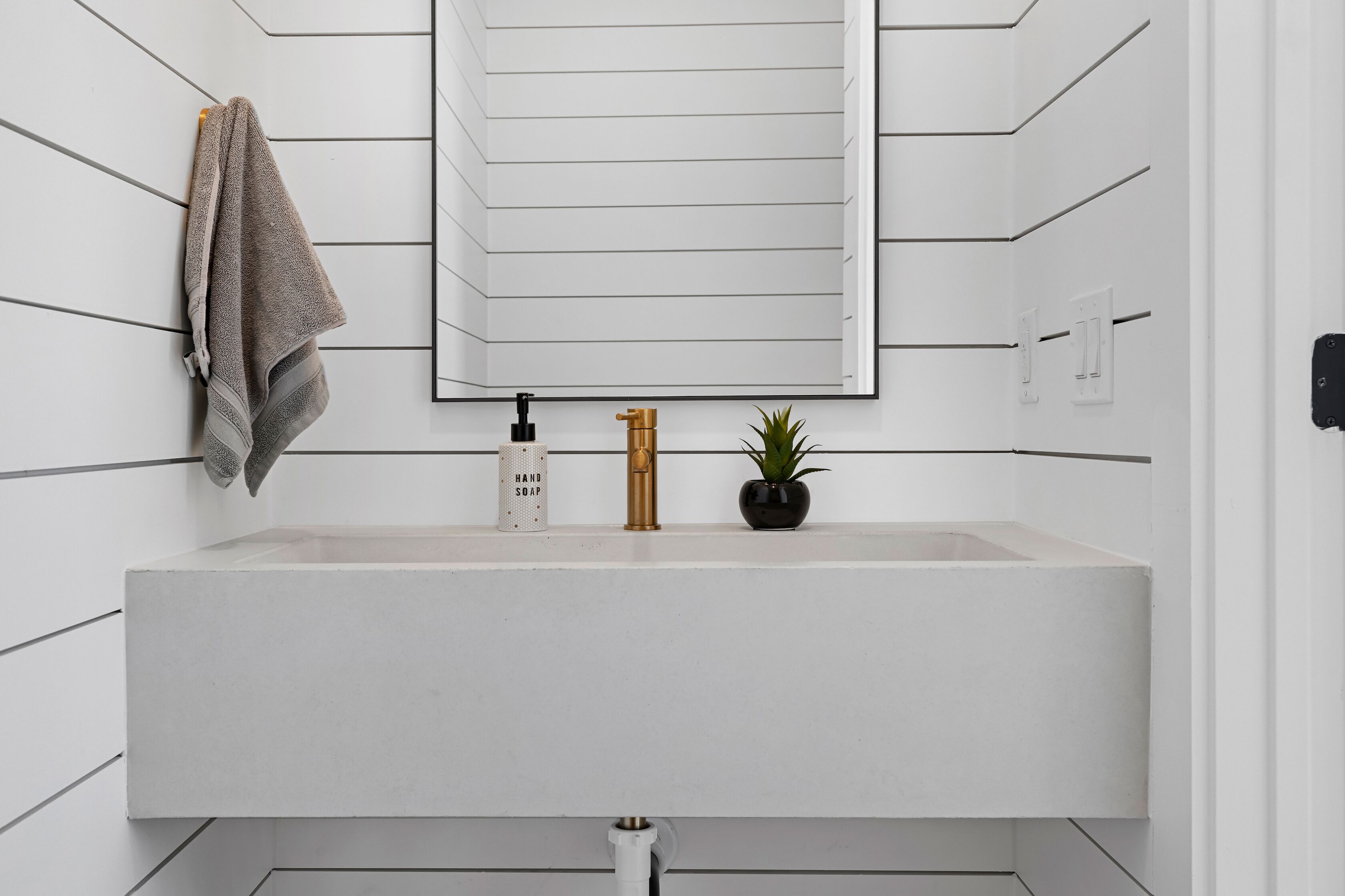 Un baño moderno con un lavamanos blanco, una toalla colgada en la pared, una pequeña planta en maceta y un espejo sobre el lavamanos.
