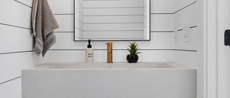 Un baño moderno con un lavamanos blanco, una toalla colgada en la pared, una pequeña planta en maceta y un espejo sobre el lavamanos.