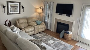 Ein Wohnzimmer mit beiger Couch, grauer Couch, Kamin, Fernseher und einem gerahmten Bild an der Wand.