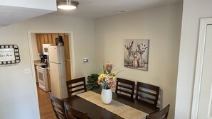 Ein Essbereich mit einem Holztisch, vier Stühlen, einer Vase mit Blumen, einem Gemälde an der Wand und einer Küche im Hintergrund.