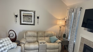 Ein Wohnzimmer mit einem beigefarbenen Ledersofa, einem gemusterten Zierkissen, einem kleinen Beistelltisch mit Lampe, einem gerahmten Bild an der Wand und einem Kamin.