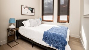 Een slaapkamer met een bed, nachtkastje, lamp, raam en een schilderij aan de muur.