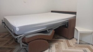 Ein Einzelzimmer mit Klappbett, einem kleinen Nachttisch und einem an der Wand befestigten Lichtschalter.