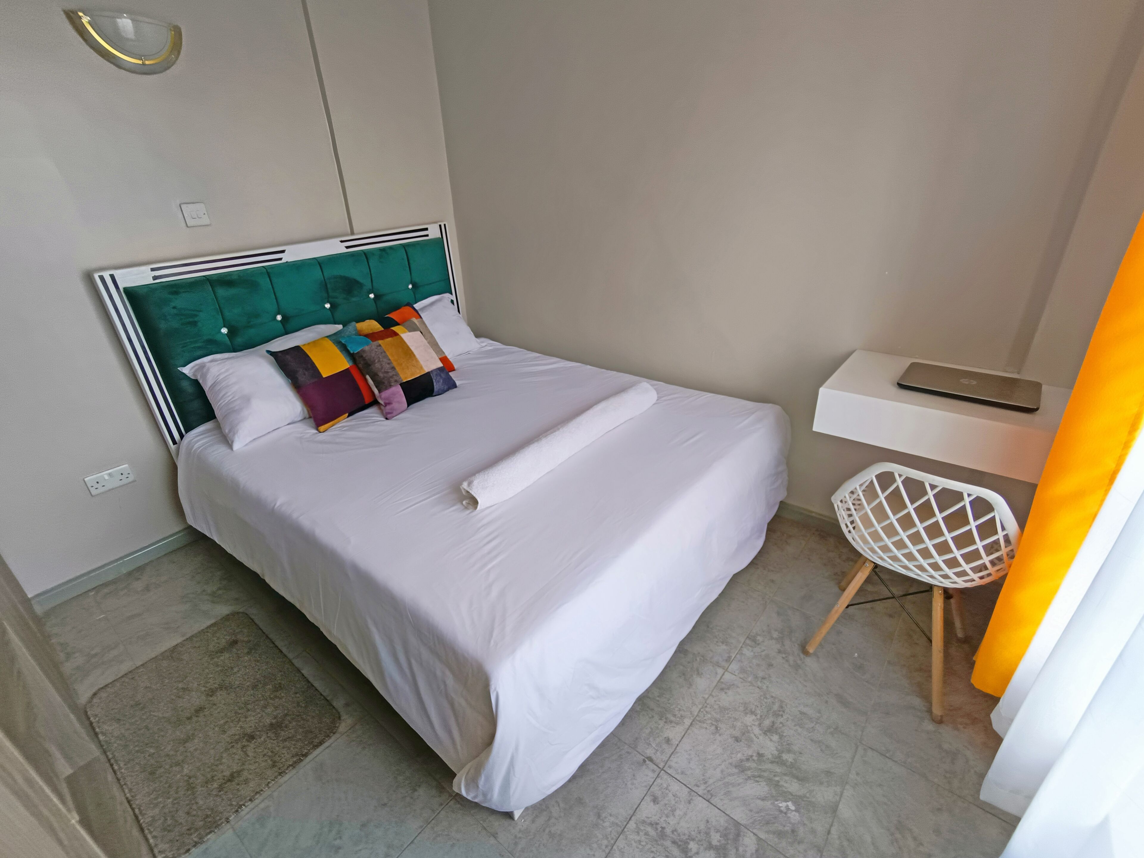 Un lit bien fait, avec une tête de lit verte, un petit bureau sur lequel se trouve un ordinateur portable et une chaise blanche.