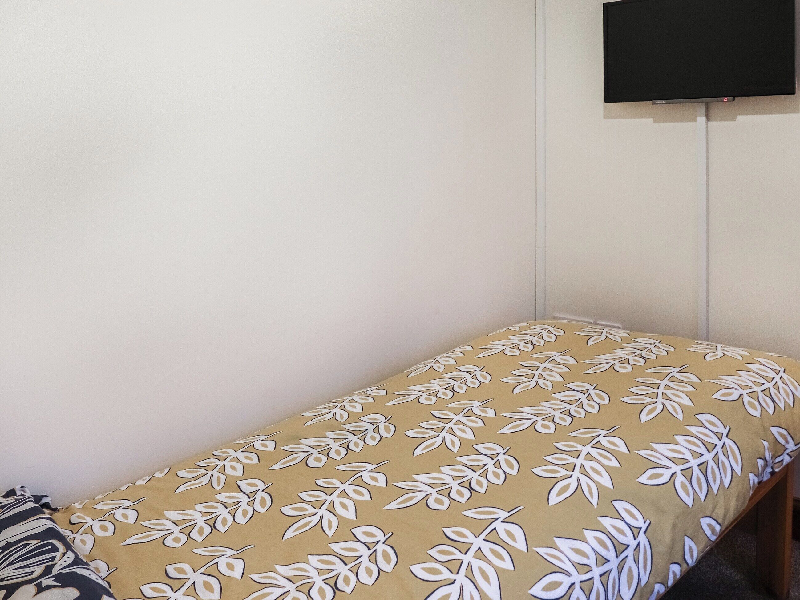 Un lit simple avec une couvre-lit à motifs, dans une pièce aux murs blancs et dotée d’un téléviseur à écran plat fixé au mur.