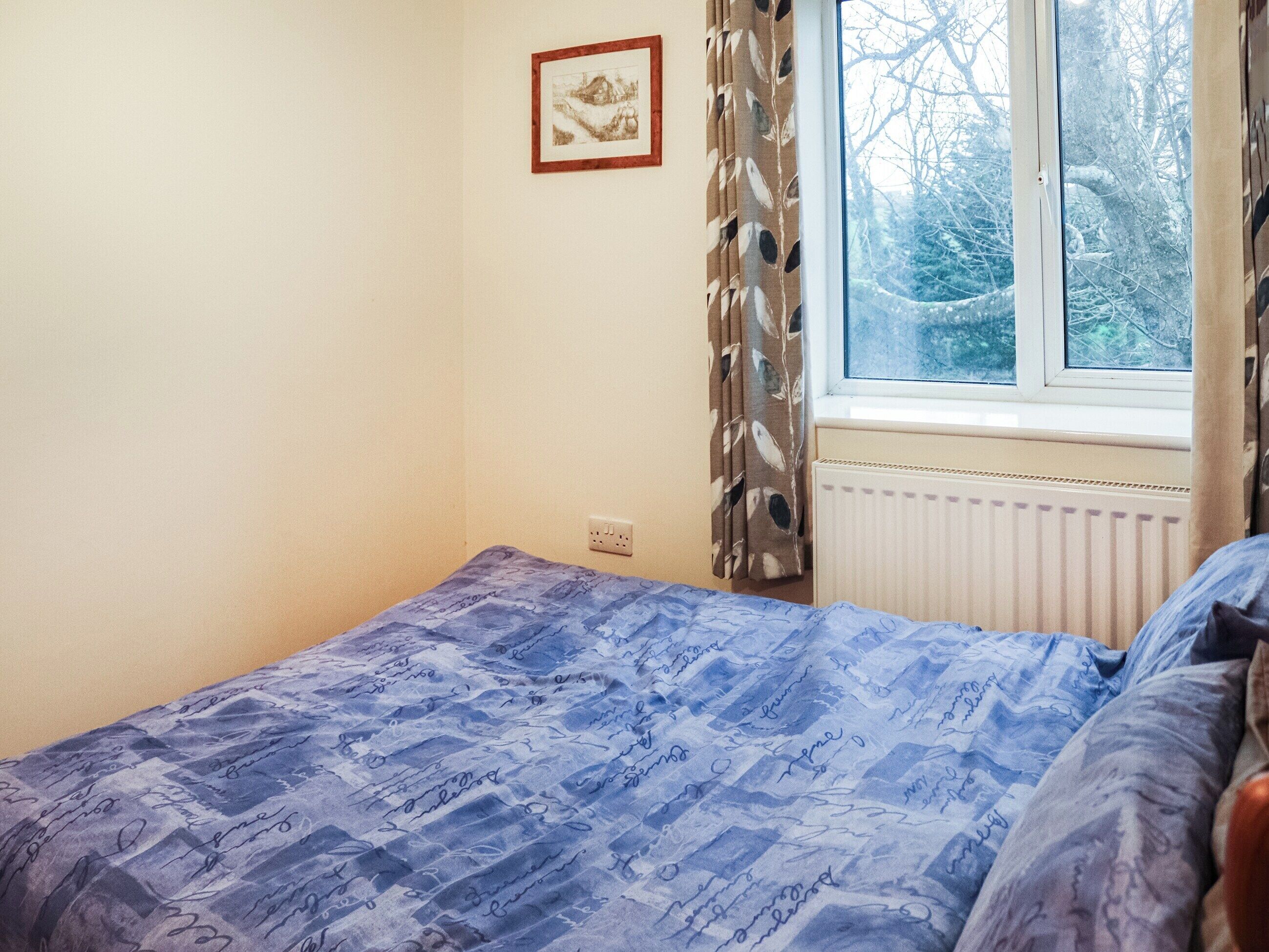 Une chambre à coucher avec un lit, une fenêtre avec des rideaux, un radiateur et un tableau encadré au mur.
