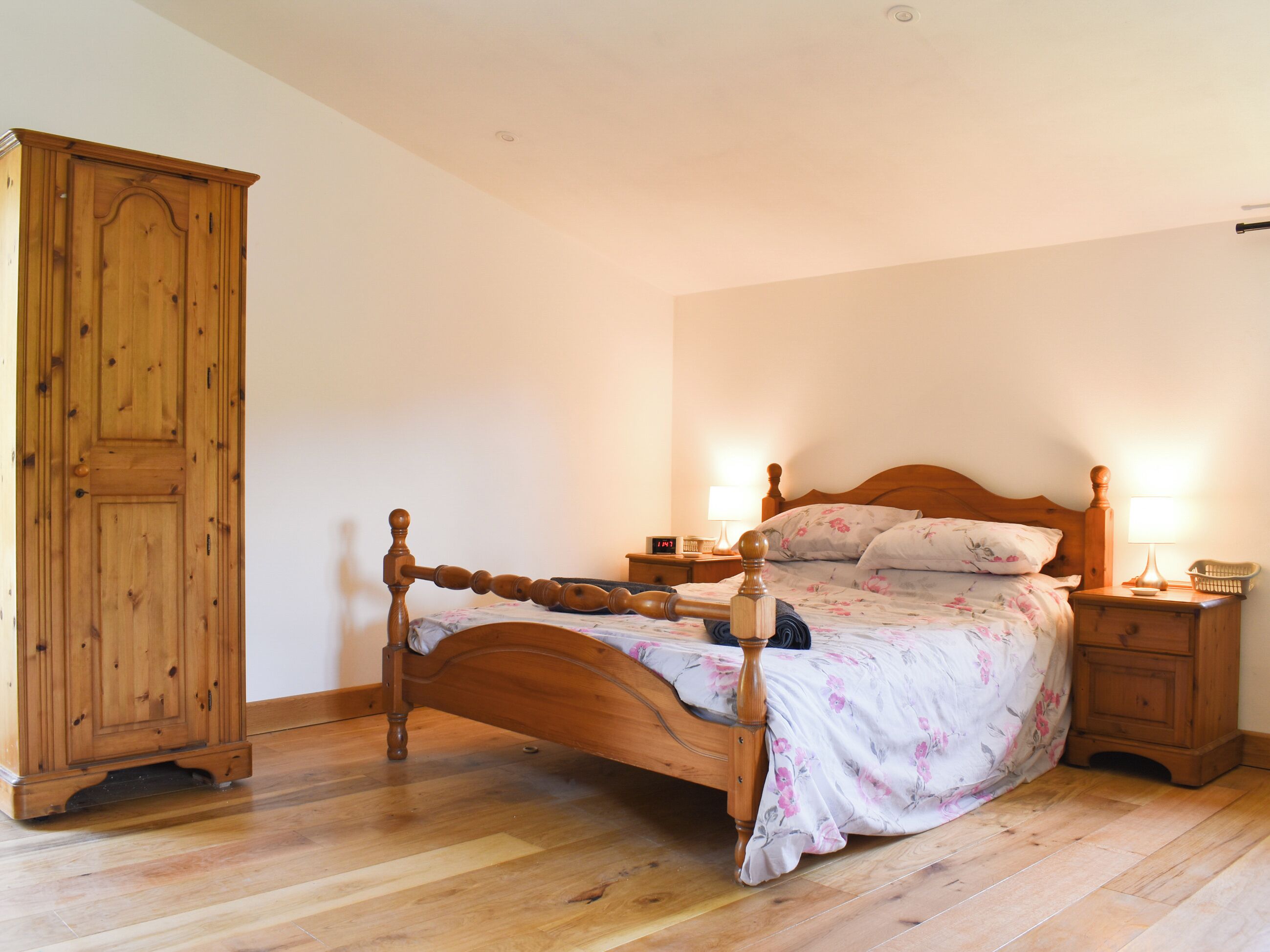 Una cama de madera con una colcha estampada, dos mesitas de noche con lámparas y un armario de madera en una habitación con suelo de madera.