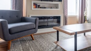 A living room with a grey sofa, a wooden coffee table, a flat-screen TV mounted on the wall, and a fireplace below. - Unmatched City View! King Bed, 2BR Apt. Free Parking! (Pittsburgh)