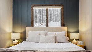 A bed with a quilted white duvet, two bedside lamps, and a window with a view of a snowy forest. - Shanty Creek Escape l Ski l Hot Tub l Pool (Bellaire)