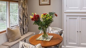 En matplats med ett runt träbord, flätade stolar, en bänk med kuddar, en vas med blommor och ett fönster med persienner och gardiner.