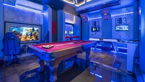 A pool table with a red felt surface, a pool cue, and a billiard ball rack. There is a large screen displaying a video game. The room has a modern design with blue lighting and a marble floor. - House Resort at Encore Reunion - Disney (Kissimmee)