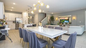 A modern kitchen with white cabinets, a stainless steel refrigerator, and a dining area with a glass table and gray chairs. - House Resort at Encore Reunion - Disney (Kissimmee)