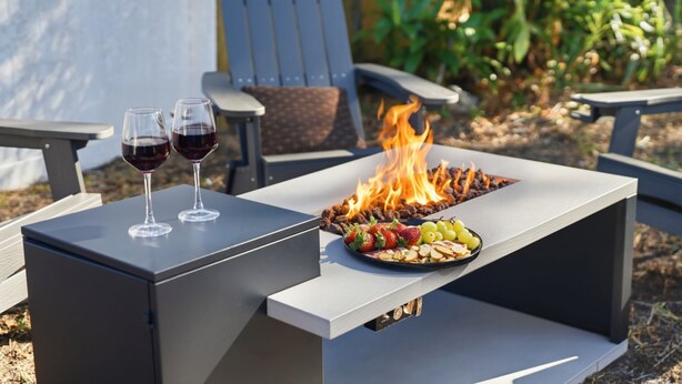 A modern outdoor grill with a fire, two glasses of red wine, and a plate of fruit on a metal table.