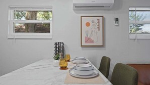 Un comedor con una mesa blanca dispuesta para cuatro personas, una obra de arte abstracta enmarcada y una ventana con persianas.