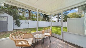 Eine überdachte Terrasse mit Korbstühlen, einem Glastisch, weißem Zaun und einem Schuppen im Hintergrund.