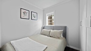 Une chambre à coucher avec un lit, deux reproductions botaniques encadrées, une fenêtre avec des stores et une porte.