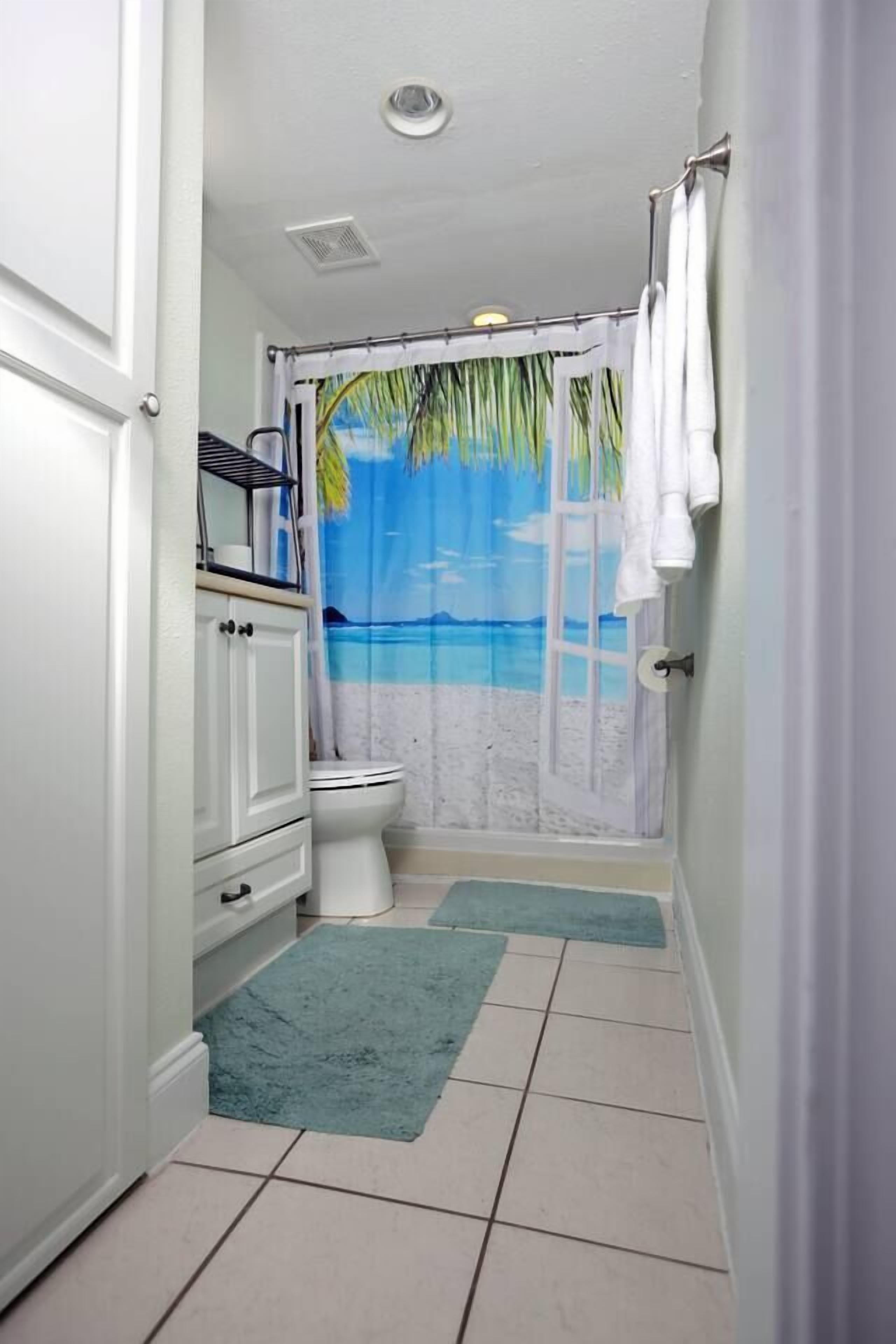 A bathroom with a toilet, a cabinet, a shower curtain depicting a beach scene, and two mats on the floor.