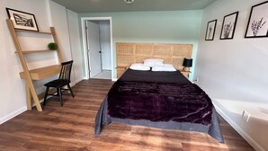 A bedroom with a bed, a chair, a desk, and a door leading to another room.