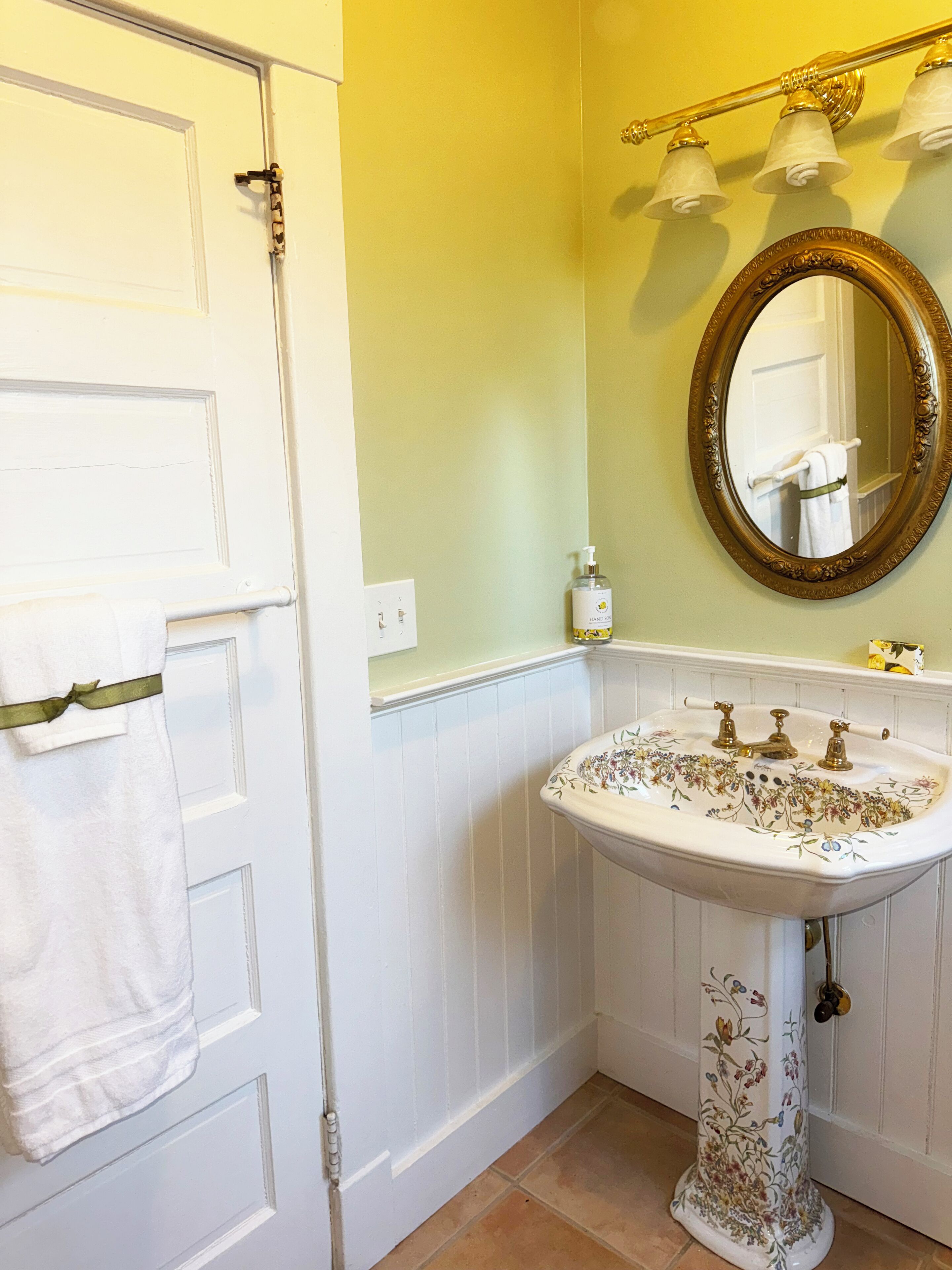 Une salle de bain avec une porte blanche, un lavabo blanc orné de motifs floraux, un miroir avec un cadre doré et un porte-serviettes agrémenté d’un ruban vert.