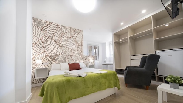 A modern bedroom with a bed, a green throw blanket, a black chair, a white bedside table, and a wall with a geometric pattern. - VIZCAINOS GARDEN (Segovia)