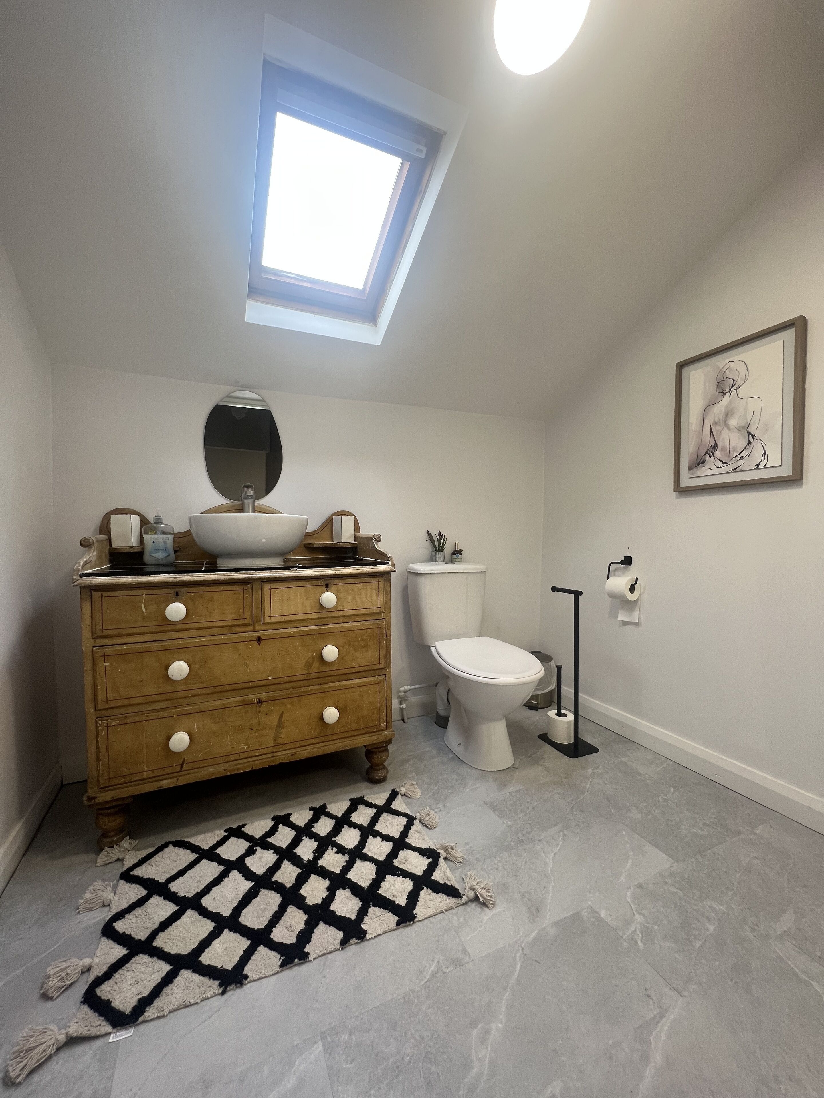 A bathroom with a skylight, a wooden dresser, a toilet, and a framed artwork on the wall.