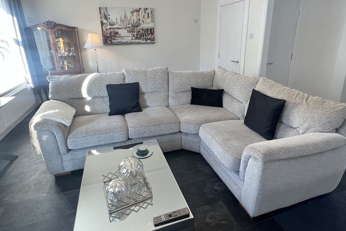 Ein modernes Wohnzimmer mit einer grauen Eckcouch, einem Glastisch, einer Kronleuchter und einem Stadtbild an der Wand.