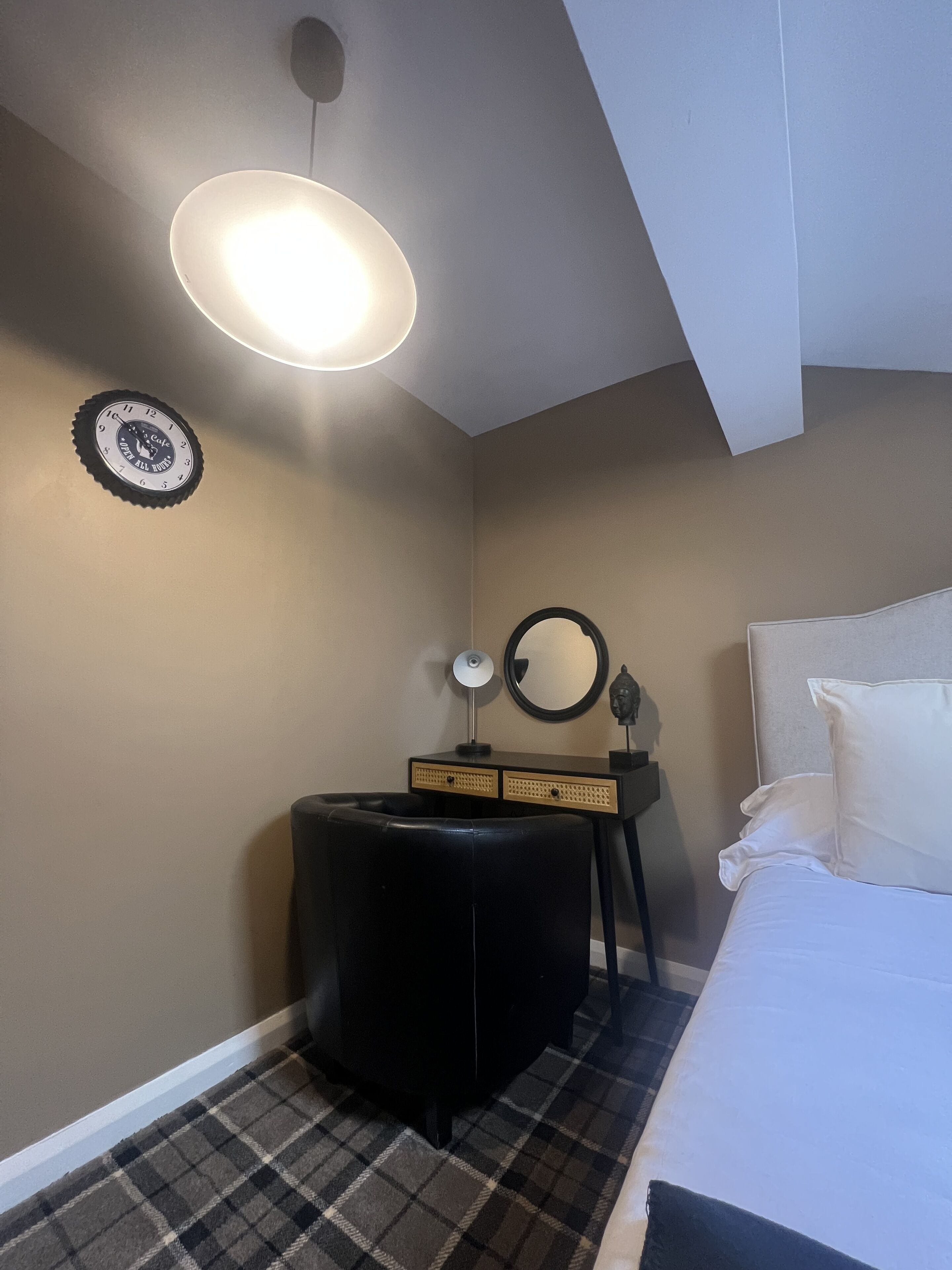 A bedroom with a bed, a black ottoman, a desk with drawers, a mirror, and a clock on the wall.